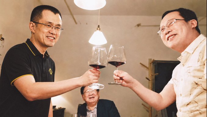 Two men toast with glasses of red wine at a gathering, while another person sits smiling in the background.