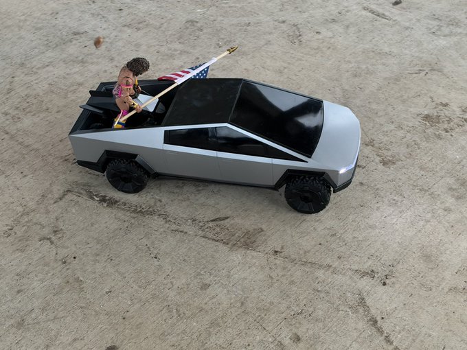A small silver toy Tesla Cybertruck sits on a concrete surface, featuring a miniature figure holding an American flag in the open rear bed.