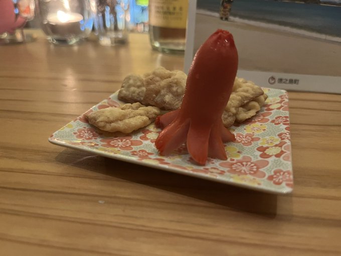 A small square plate with cherry blossom designs holding what appears to be a tomato carved to resemble a squid, served with crackers at a restaurant.