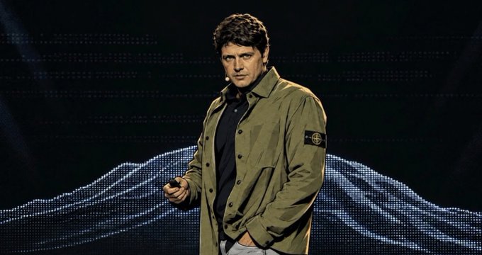A man wearing a Stone Island jacket stands on a dimly lit stage with a wavy digital display behind him, appearing to deliver a keynote presentation—likely the "const" mentioned in the post who "absolutely cooked with that opening speech" at the Exploit Summit, discussing progress around $TAO and subnet developments.