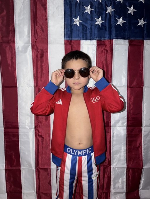 A young boy wearing an open red Olympic Team USA jacket, red, white, and blue Olympic pants, and aviator sunglasses, posing in front of an American flag.