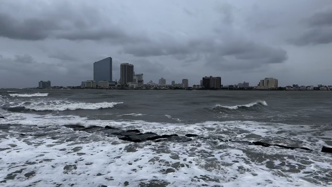 A stormy, overcast coastal scene showing rough waves crashing against rocks in the foreground with a city skyline of high-rise buildings visible across the water.