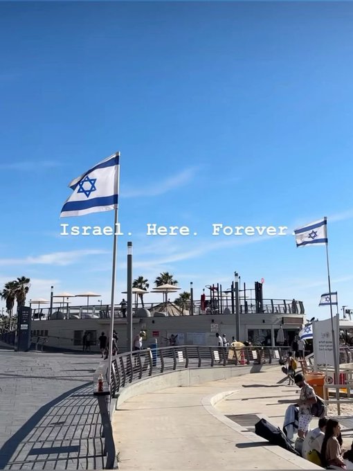 A photograph of a coastal promenade with Israeli flags waving and the text "Israel. Here. Forever" overlaid across a clear blue sky.