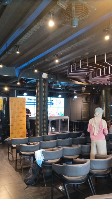 The image shows a room with several chairs arranged in rows, some of which are empty while others have backpacks placed on them. A mannequin wearing a pink shirt stands near one of the rows of chairs, possibly for display or as part of an event setup. The room also features a large screen and a banner hanging from the ceiling, adding to its ambiance. The post in the image is related to Bitcoin and seems to be promoting an event at the Binance Academy.