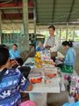 A small, low-resolution photo showing people working at what appears to be a market or food preparation stall under a sheltered area.