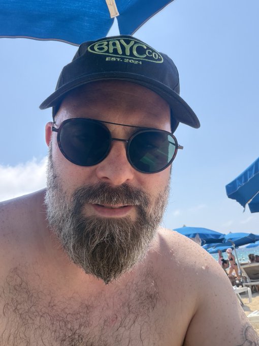A shirtless bearded man takes a beach selfie wearing a black cap emblazoned with "BAYC CO EST. 2021," which ties directly to the "BAYC CO 4L" reference—signaling his allegiance to the Bored Ape Yacht Club community under sunny seaside conditions.
