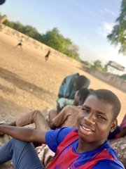 A young man in a red and blue striped shirt smiles at the camera while sitting outdoors with other people in what appears to be a dry, open area.