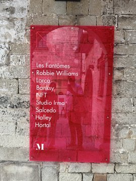 A red exhibition poster on a stone wall listing artists including Les Fantômes, Robbie Williams, Loco, Banksy, NFT, Studio Irma, Salcedo, and Hailey Hortal.