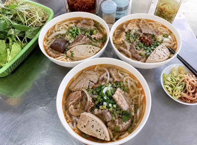 Three bowls of Vietnamese bún (beef noodle soup) topped with sliced beef, pork, and meatball, garnished with green onions and chili sauce, served with a side of fresh herbs and bean sprouts. The image showcases a hearty breakfast spread that aligns with the post's mention of treating his wife to a free, well-earned meal to enjoy the flavorful noodle bowls together.