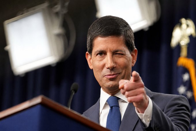 A man in a dark suit and blue tie pointing forward while standing at a podium with a US flag in the background.