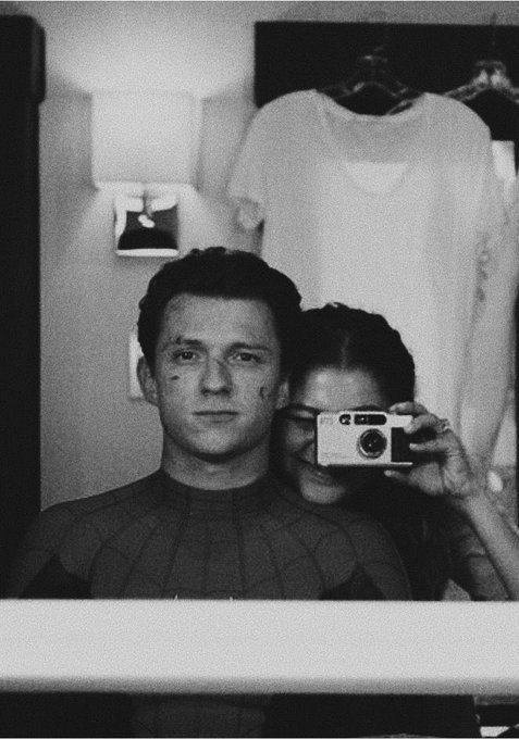 A black-and-white mirror selfie of actor Tom Holland in a Spider-Man suit, with actress Zendaya partially visible behind him holding a camera, taken in what appears to be a dressing room.