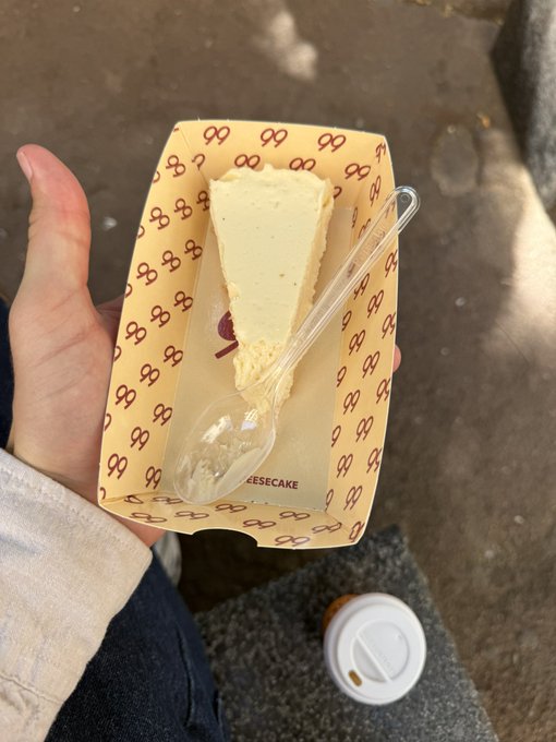 A hand holding a small paper tray containing a slice of cheesecake with a clear plastic spoon, taken outdoors on pavement with a coffee cup lid visible below.