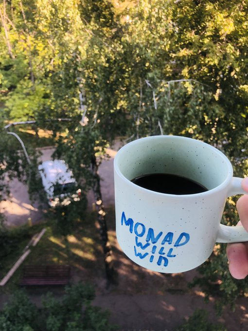 A hand holding a speckled light blue ceramic mug filled with dark coffee over a balcony with a blurred background of green trees and a park below; the mug has 'MONAD WILL WIN' written on it in blue marker.