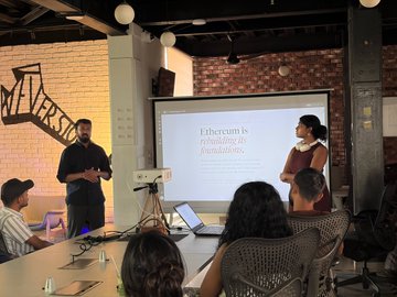 A presentation/talk scene where a man in black stands near a screen displaying 'Ethereum is rethinking its foundations' while a woman in a red dress speaks, with an audience seated at tables in what appears to be a meetup venue.