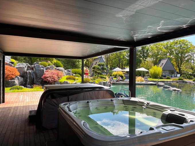A luxurious outdoor relaxation area featuring a hot tub under a covered patio, overlooking a landscaped garden with a pond, Japanese-style architecture, and lounge seating in the background.
