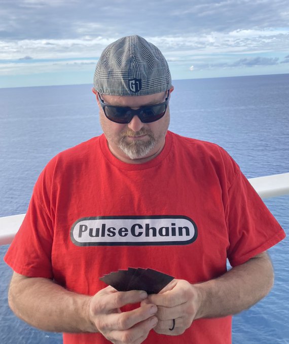A man wearing a red 'PulseChain' t-shirt, sunglasses, and a grey flat cap, standing outdoors against a blue sky and white railing while holding what appears to be trading cards.