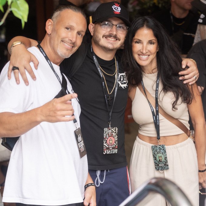 Three people pose together at what appears to be an event, with the young man in the center wearing a black cap, glasses, and an event lanyard, flanked by a smiling older man in white and a woman in a cream-colored outfit.