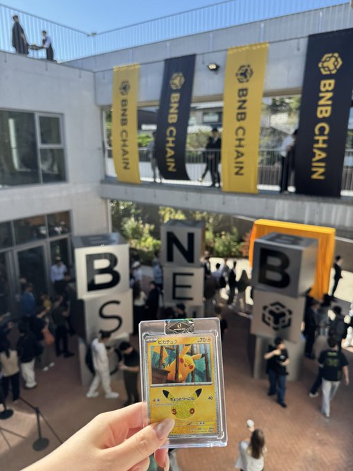 A hand holding a Japanese Pokémon card (Pikachu) at what appears to be a BNB Chain event, with large 'BNB CHAIN' banners and block letter sculptures in the background.