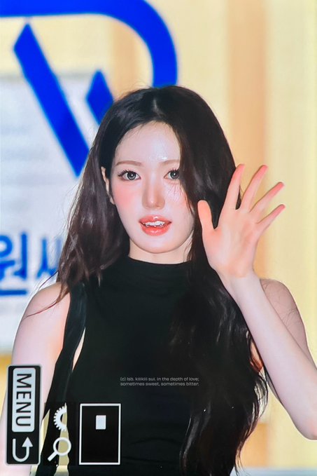 A woman with long dark hair wearing a black sleeveless top waves her hand, with text overlay reading "(ɔ) laï, kïssïï süï, in the depth of løvë: sometimes sweet, sometimes bitten" and a camera menu UI at the bottom.