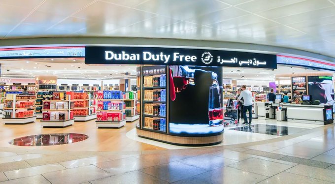 The interior of a 'Dubar Duty Free' store, featuring a central display with large digital screens showing red liquid being poured, with various product shelves and a customer browsing in the background.