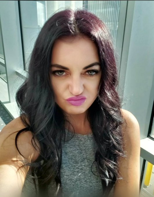 A selfie of a woman with dark purple hair and pink lipstick making a pouting expression, wearing a gray tank top.