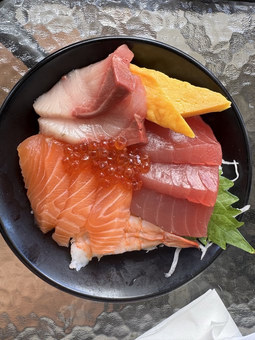 A black bowl filled with a colorful assortment of fresh sashimi, including salmon, tuna, yellowtail, shrimp, and salmon roe, garnished with a shiso leaf and daikon.