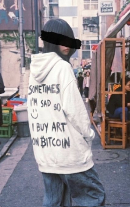 The image features a person standing on a street, wearing a hoodie with graffiti writing on it that reads "Sometimes I'm sad so I buy art on Bitcoin". The person's face is obscured by a black bandana covering their eyes. In the background, there are several other people walking around or sitting at tables outside of a building.