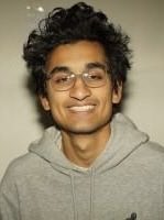 A smiling young man with curly black hair and glasses, wearing a gray hoodie, looks confidently at the camera, embodying a determined and resilient spirit that matches the caption "This Indian doesn't lose."