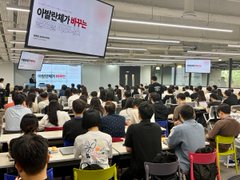A large group of people sits at tables in a brightly lit room facing a screen displaying Korean text, suggesting an educational or training session.