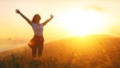 A woman stands in a field with her arms raised in celebration against a bright golden sunset.