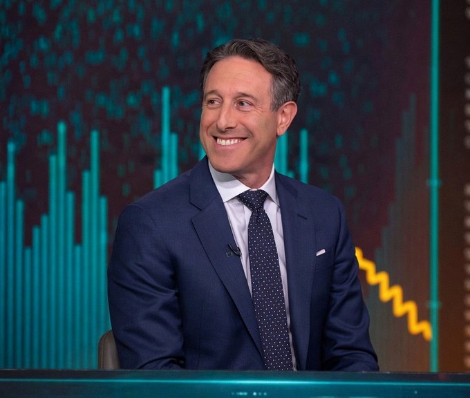 A smiling man in a navy suit and dotted tie, likely a television host, seated in front of a teal financial-style graphic.