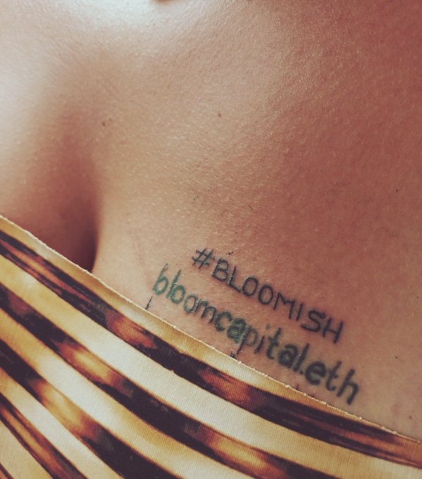 A close-up photo showing a tattoo on a person's chest/collarbone area with the text '#BLOOMISH' and 'bloomcapital.eth' inked in black, visible just above a patterned yellow garment.