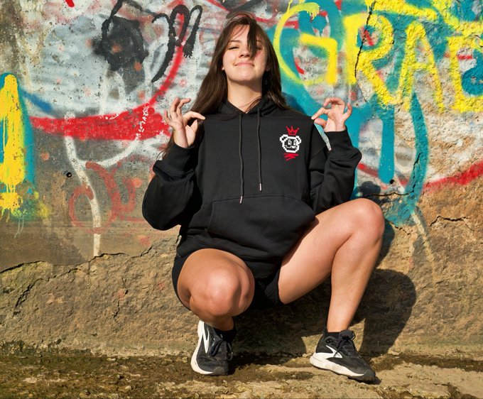 A smiling young woman crouches in front of a colorful graffiti wall, wearing a black hoodie and shorts, making finger heart gestures with both hands.