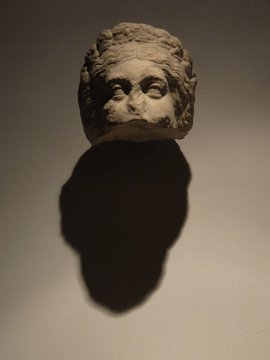 Stone sculpted head mounted on a wall, casting a strong shadow against a plain background.