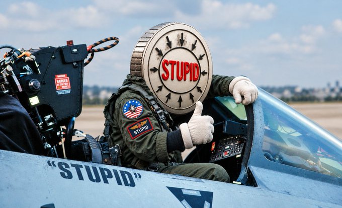 A pilot in a fighter jet cockpit wears a helmet with 'STUPID' written in red letters on a clock-like face, while the jet itself is labeled 'STUPID' on the side, and the pilot gives a thumbs-up gesture.