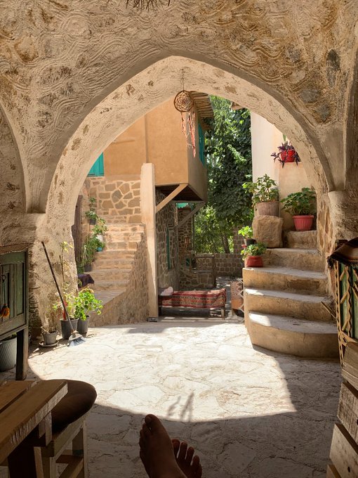 A scenic view through a stone archway of a sunlit courtyard with stone steps, potted plants, and a traditional rug in a rustic stone building.