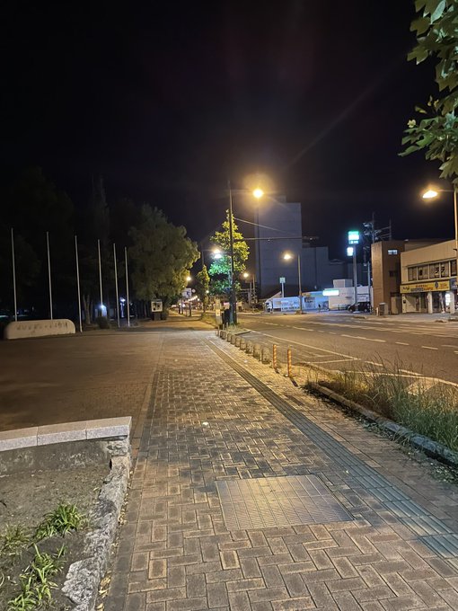 The image shows a city street at night with a brick sidewalk, lampposts, and buildings lining both sides of the road. The scene appears to be empty or quiet, as there are no people visible in the photo. A caption on the image reads "めっちゃ台風なんですけど、とりあえず傘持ってこれから出かけます。写真は普段の定例深夜コースです".