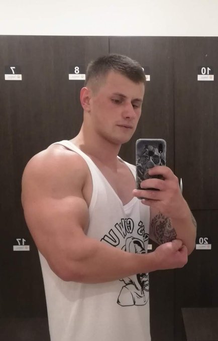 Mirror selfie of a muscular young man in a white tank top flexing his bicep in a locker room, wearing a phone case with a skeleton design.