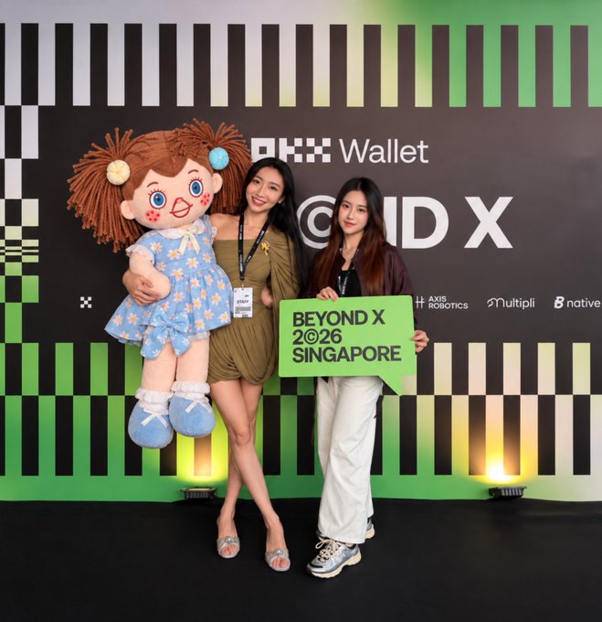 Two women pose at the "BEYOND X 2026 SINGAPORE" event, with one holding a giant plush doll of "Paula" in a blue star-patterned dress. The backdrop features logos including "OKX Wallet," "Multipli," "AXIS ROBOTICS," and "native," indicating this is the OKX-sponsored event the poster missed in person. The accompanying post references Paula (the doll), Token2049/OKX, and tagging @PolarCoinXlayer and @EudemoniaCC, suggesting the photo was shared by an attendee on behalf of the post's author.