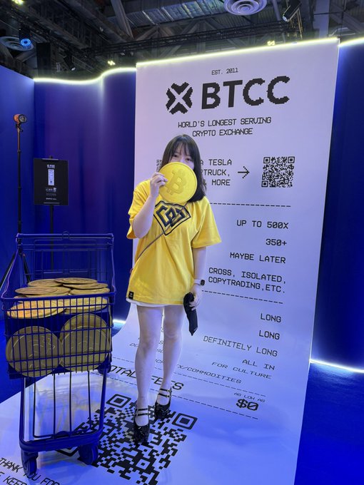 A woman in a yellow t-shirt and white skirt holds a Bitcoin token in front of a BTCC display at a crypto exchange booth.