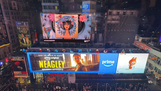 An aerial nighttime view of Times Square in New York City shows digital billboards, including an NFT display featuring a flower-crowned woman, an Amazon Prime advertisement for the series 'NEAGLEY', and the '1540 Broadway' sign.