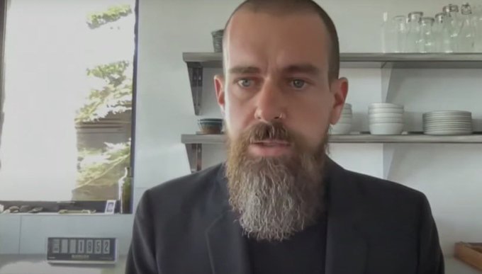 A man with a long gray beard in a dark blazer sits in what appears to be a kitchen or café, with a digital clock and dishware visible behind him.