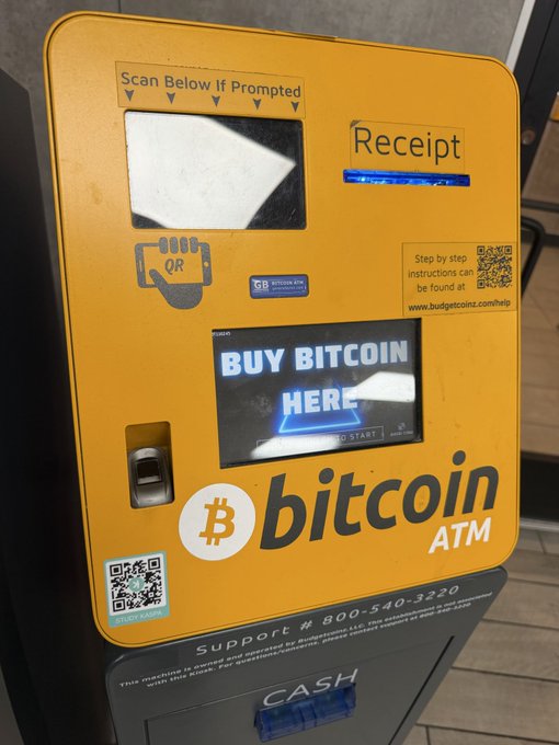 A yellow Bitcoin ATM with a screen reading 'BUY BITCOIN HERE', featuring a QR code, support number '800-540-3230', and a cash deposit slot at the bottom.