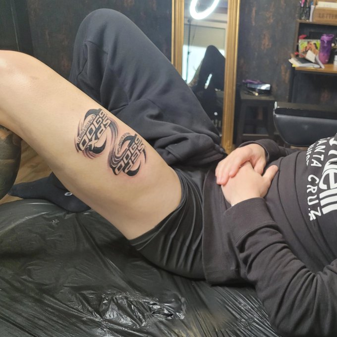 A person lies on a tattoo chair displaying two fresh SPX logos on their calf—the original "SPX Sports" design and a stylized "Salinas Pacific Express" variation—with the artist wearing a "Santa Cruz" hoodie in the background, humorously illustrating the ongoing debate over which SPX logo should be the official one.