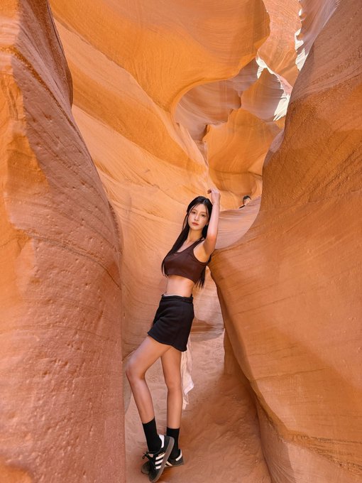 A young woman posing in a narrow slot canyon with smooth, orange sandstone walls; she wears a brown top, black skirt, and boots.