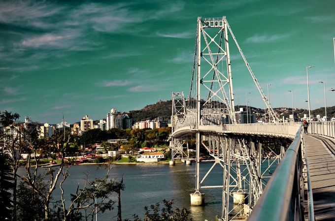 A scenic view of a large suspension bridge (Hercílio Luz Bridge) spanning a body of water with a city and hills in the background.
