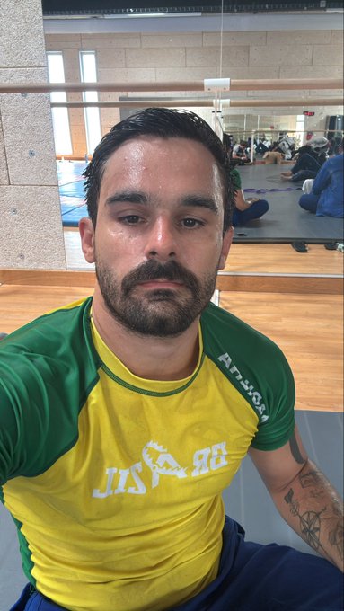 A sweaty man in a yellow and green Brazil "Jogar" shirt takes a selfie in what appears to be a martial arts dojo, with other people visible on mats in the background.