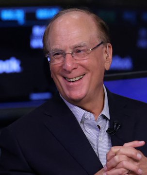 A man with glasses and a receding hairline smiles broadly while wearing a dark suit jacket over a light blue collared shirt.
