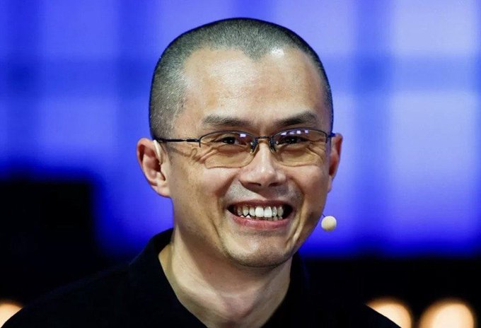 A smiling bald man wearing glasses and a black shirt speaks into a small lapel microphone against a blue backdrop, appearing to be giving a presentation or interview. The image relates to the post about Binance CEO Changpeng Zhao (CZ) discussing Bitcoin's deflationary nature and the significant portion of mined BTC that has been lost or become unrecoverable.