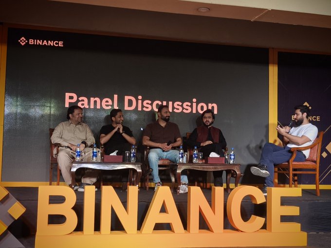 A panel discussion at a Binance event featuring five men seated on stage, with a large 'BINANCE' sign in the foreground and 'Panel Discussion' displayed on a screen behind them.